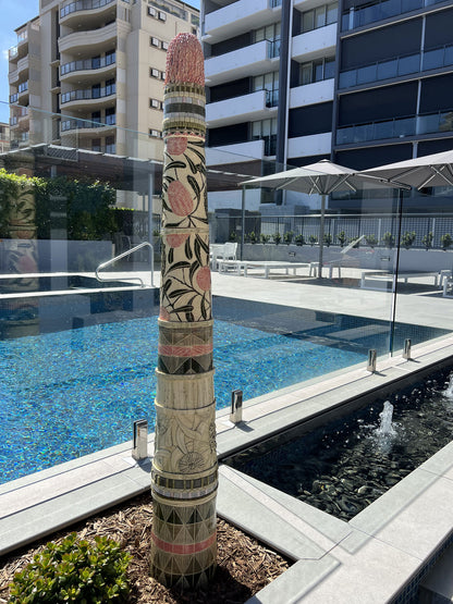 Tall ceramic sculpture with pink banksia flowers and mosaic inlay displayed on a balcony beside a swimming pool